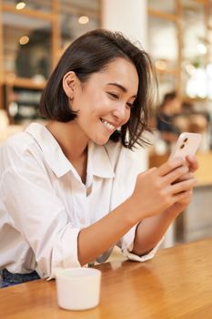 Vertical shot of brunette asian girl sitting in cafe, using smartphone app, chatting on mobile phone and smiling.