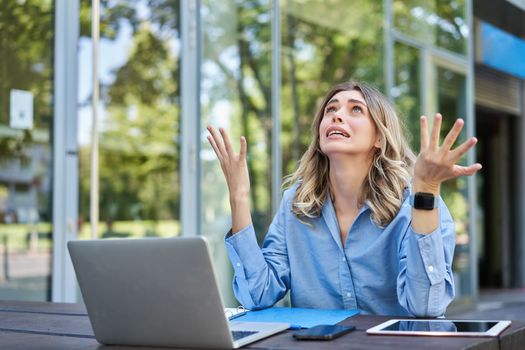 Disappointed young businesswoman feeling frustrated, sitting with laptop outdoors on street, looking up and complaining.