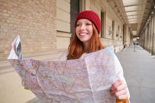 Smiling young redhead woman in red hat, looks at paper map to look for tourist attraction. Tourism and people concept. Girl explores city, tried to find way.