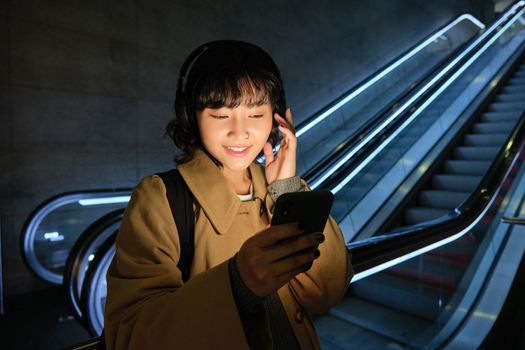 Young girl in headphones looks at her mobile phone, stands near escalator, listens to music while travels in city.
