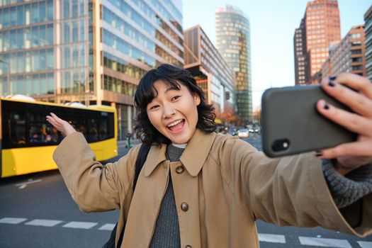 Upbeat asian girl takes selfie with smartphone in city centre, showing something behind her with smiling face. Tourist goes sightseeing.