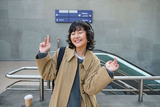 Happy young girl, student has fun on street, dances, listens music in headphones, drinks coffee. Copy space