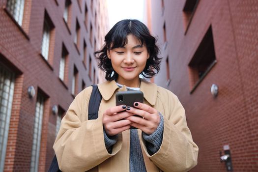 Lower angle view of brunette korean girl, listening music in headphones, walking along street and looking at smartphone, reading message on mobile phone.