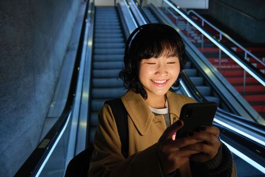 Portrait of girl student in headphones, listens music, commutes, goes down escalator, looks at smartphone with pleased smile, happy face.