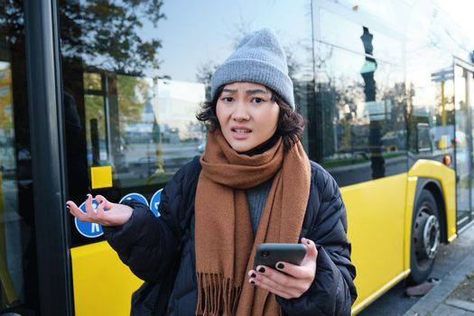 Portrait of confused young woman, looking at her smartphone app with shocked and disappointed face, shrugs shoulders, reads bad news, stands on bus stop near public transport.
