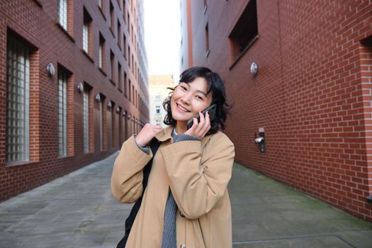 Portrait of young korean woman walking down street with phone, talking with someone, makes a call, has telephone conversation.