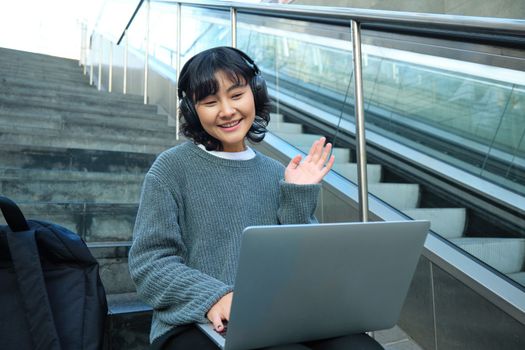 Smiling young student, wears headphones, sits on street stairs and waves hand at her laptop, connects to video chat, conference or training session on remote.