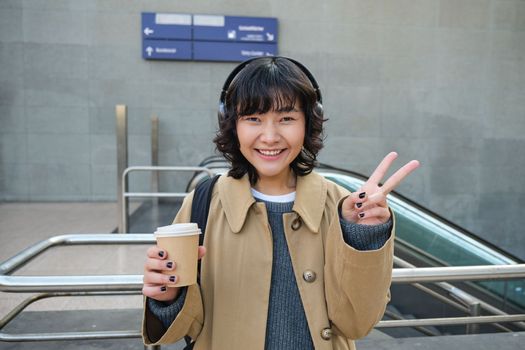 Positive korean girl in headphones, drinks coffee to go, shows peace, v-sign, stands on street and smiles happily.