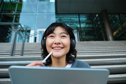 Close up of smiling asian girl feels inspired while draws on digital tablet, makes graphic design project, pencils user interface assignment, sits on stairs near campus.