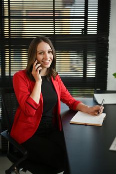 Portrait of smiling caucasian female manager in elegant red suit having pleasant conversation on mobile phone.