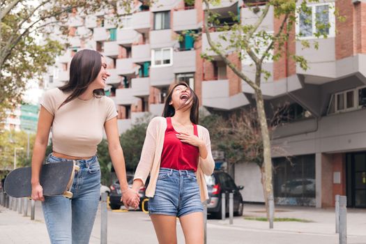 couple of women friends walking and laughing holding hands through the city, concept of female friendship and love