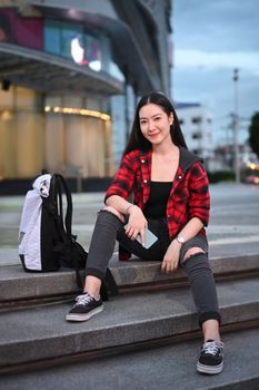 Portrait of young Asian woman sitting on stairs in city at night and smiling to camera.