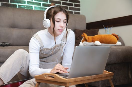 A young female student sits at a table and studies in headphones. A girl in headphones watches a webinar, listens to an online course while communicating on a conference video call.