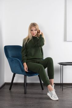Stylish beautiful young blond woman in a green tracksuit poses near a white wall in the room. Attractive girl model posing in a blue chair. Fitness lady
