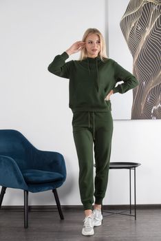 Stylish beautiful young blond woman in a green tracksuit poses near a white wall in the room. Attractive girl model posing near blue chair. Fitness lady
