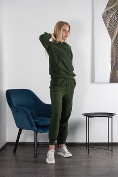 Stylish beautiful young blond woman in a green tracksuit poses near a white wall in the room. Attractive girl model posing near blue chair. Fitness lady