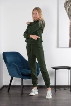 Stylish beautiful young blond woman in a green tracksuit poses near a white wall in the room. Attractive girl model posing near blue chair. Fitness lady