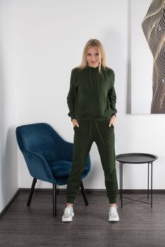 Stylish beautiful young blond woman in a green tracksuit poses near a white wall in the room. Attractive girl model posing near blue chair. Fitness lady