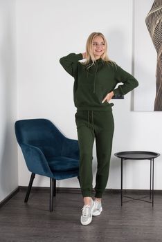 Stylish beautiful young blond woman in a green tracksuit poses near a white wall in the room. Attractive girl model posing near blue chair. Fitness lady