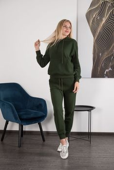 Stylish beautiful young blond woman in a green tracksuit poses near a white wall in the room. Attractive girl model posing near blue chair. Fitness lady