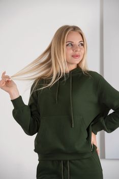 Stylish beautiful young blond woman in a green tracksuit poses near a white wall in the room. Attractive girl model posing near blue chair. Fitness lady