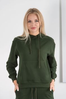 Stylish beautiful young blond woman in a green tracksuit poses near a white wall in the room. Fitness lady