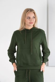 Stylish beautiful young blond woman in a green tracksuit poses near a white wall in the room. Fitness lady