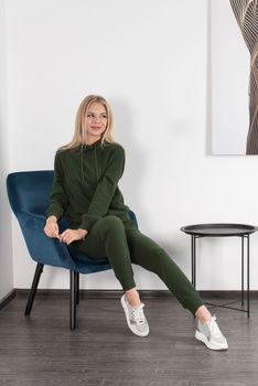 Stylish beautiful young blond woman in a green tracksuit poses near a white wall in the room. Attractive girl model posing in a blue chair. Fitness lady