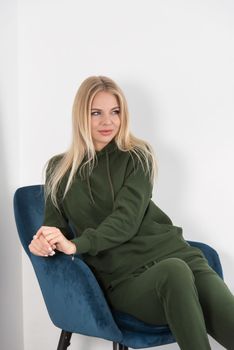 Stylish beautiful young blond woman in a green tracksuit poses near a white wall in the room. Attractive girl model posing in a blue chair. Fitness lady