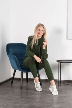 Stylish beautiful young blond woman in a green tracksuit poses near a white wall in the room. Attractive girl model posing in a blue chair. Fitness lady