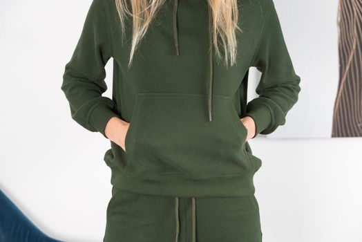 Stylish beautiful young blond woman in a green tracksuit poses near a white wall in the room. Fitness lady