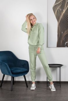 Stylish beautiful young blond woman in a light green tracksuit poses near a white wall in the room. Attractive girl model posing near blue chair. Fitness lady