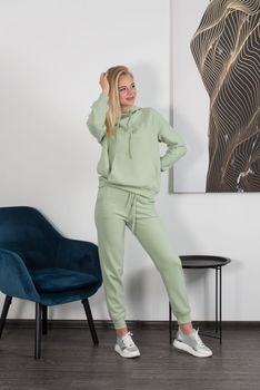 Stylish beautiful young blond woman in a light green tracksuit poses near a white wall in the room. Attractive girl model posing near blue chair. Fitness lady