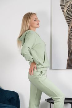 Stylish beautiful young blond woman in a light green tracksuit poses near a white wall in the room. Attractive girl model posing near blue chair. Fitness lady