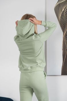 Stylish beautiful young blond woman in a green tracksuit poses near a white wall in the room. Fitness lady