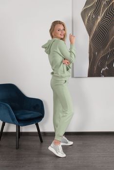 Stylish beautiful young blond woman in a light green tracksuit poses near a white wall in the room. Attractive girl model posing near blue chair. Fitness lady