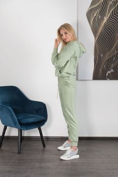 Stylish beautiful young blond woman in a light green tracksuit poses near a white wall in the room. Attractive girl model posing near blue chair. Fitness lady