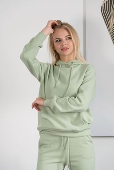 Stylish beautiful young blond woman in a green tracksuit poses near a white wall in the room. Fitness lady