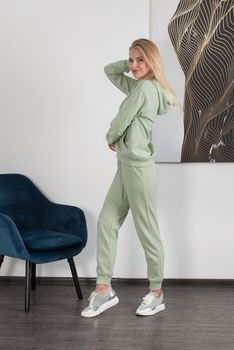 Stylish beautiful young blond woman in a light green tracksuit poses near a white wall in the room. Attractive girl model posing near blue chair. Fitness lady