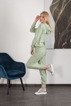 Stylish beautiful young blond woman in a light green tracksuit poses near a white wall in the room. Attractive girl model posing near blue chair. Fitness lady