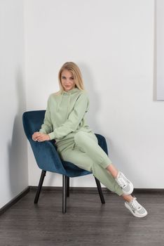 Stylish beautiful young blond woman in a light green tracksuit poses near a white wall in the room. Attractive girl model posing in a blue chair. Fitness lady