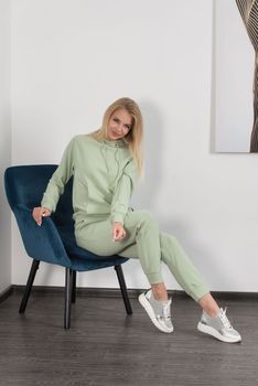 Stylish beautiful young blond woman in a light green tracksuit poses near a white wall in the room. Attractive girl model posing in a blue chair. Fitness lady