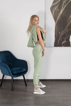 Stylish beautiful young blond woman in a light green tracksuit poses near a white wall in the room. Attractive girl model posing near blue chair. Fitness lady