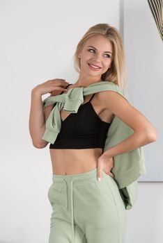 Stylish beautiful young blond woman in a green tracksuit poses near a white wall in the room. Fitness lady