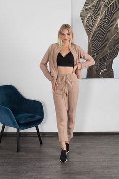 Stylish beautiful young blond woman in a biege tracksuit and black top poses near a white wall in the room. Fitness lady