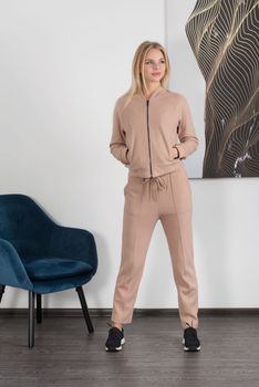 Stylish beautiful young blond woman in a biege tracksuit poses near a white wall in the room. Fitness lady