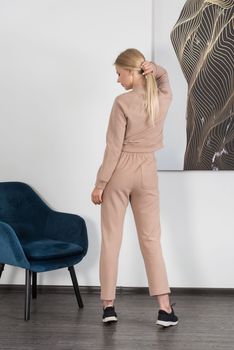 Stylish beautiful young blond woman in a biege tracksuit poses near a white wall in the room. Fitness lady