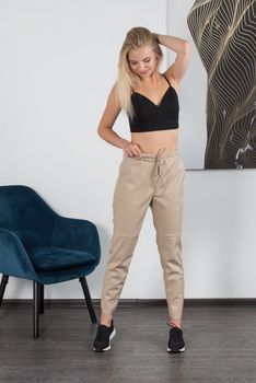 Stylish beautiful young blond woman in a tight biege pants, black top and white shirt near a white wall in the room. Attractive girl model posing near blue chair. Fitness lady