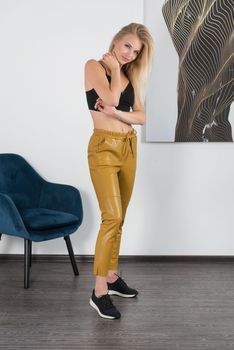 Stylish beautiful young blond woman in a tight yellow pants, black top and white shirt near blue chair in the room. Attractive girl model posing near blue chair. Fitness lady