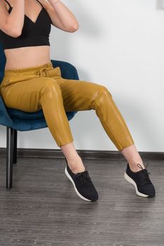 Stylish beautiful young blond woman in a tight yellow pants, black top and white shirt near a white wall in the room. Attractive girl model posing near blue chair. Fitness lady